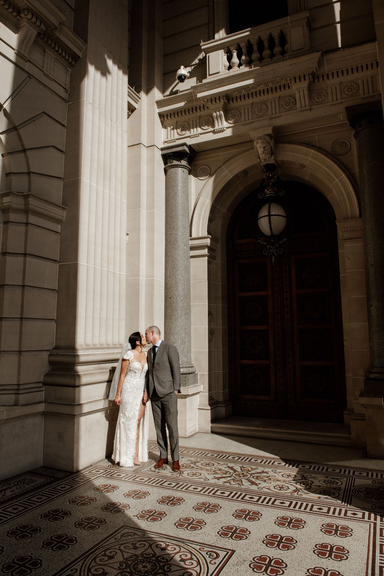 A wedding couple poses against ornate architectural columns and patterned floor in classic building.