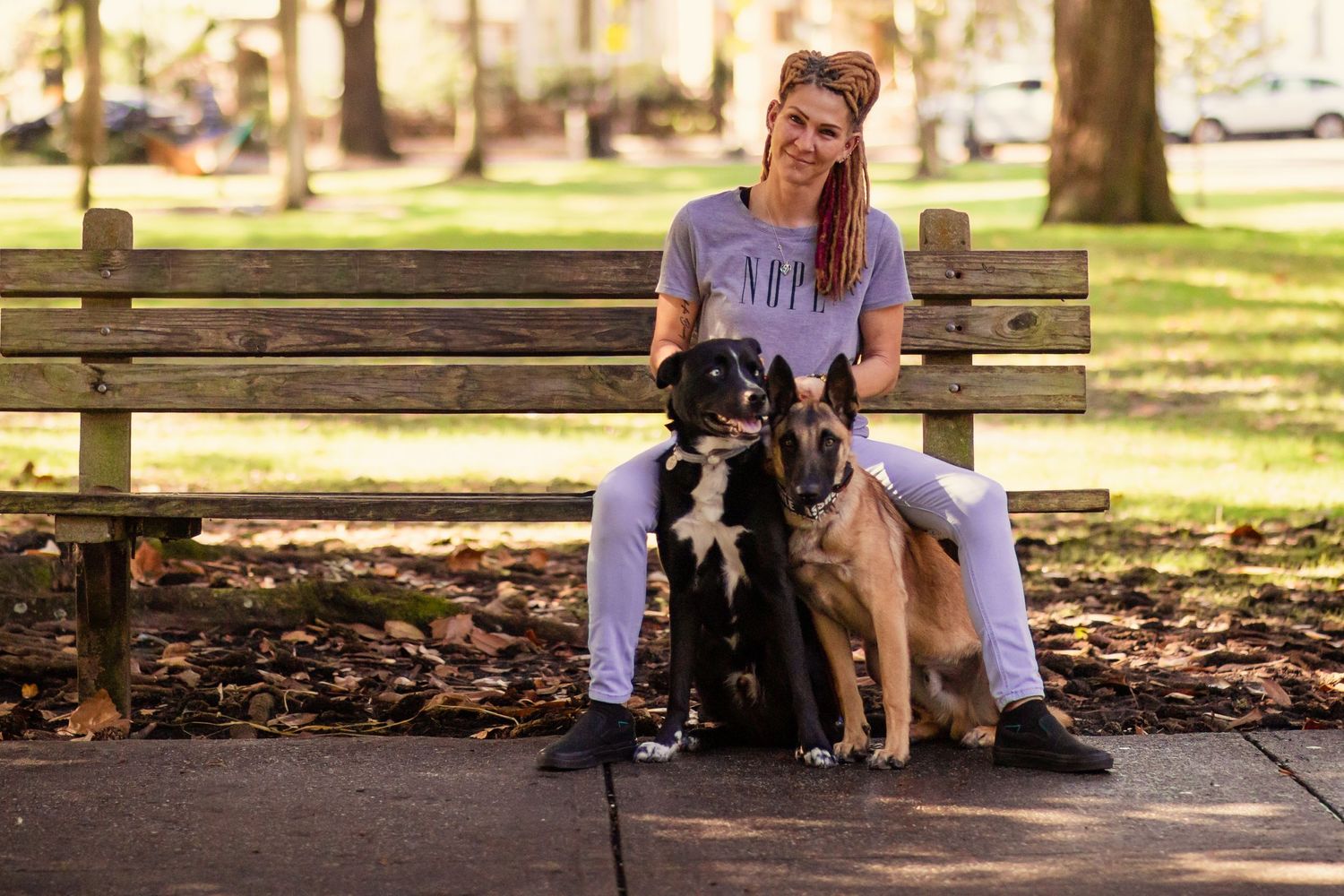 Woman with pink hair sits on bench with two dogs in a sunny park.