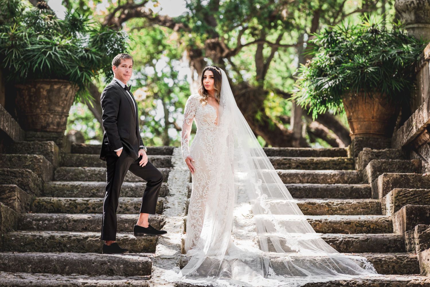 A couple poses for wedding photos on ancient stone steps surrounded by greenery.