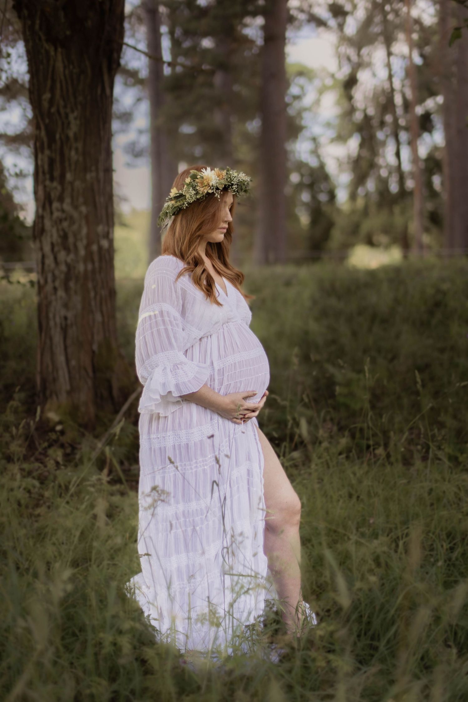 Pregnant woman wearing a floral crown in a white boho dress standing in a lush green forest.