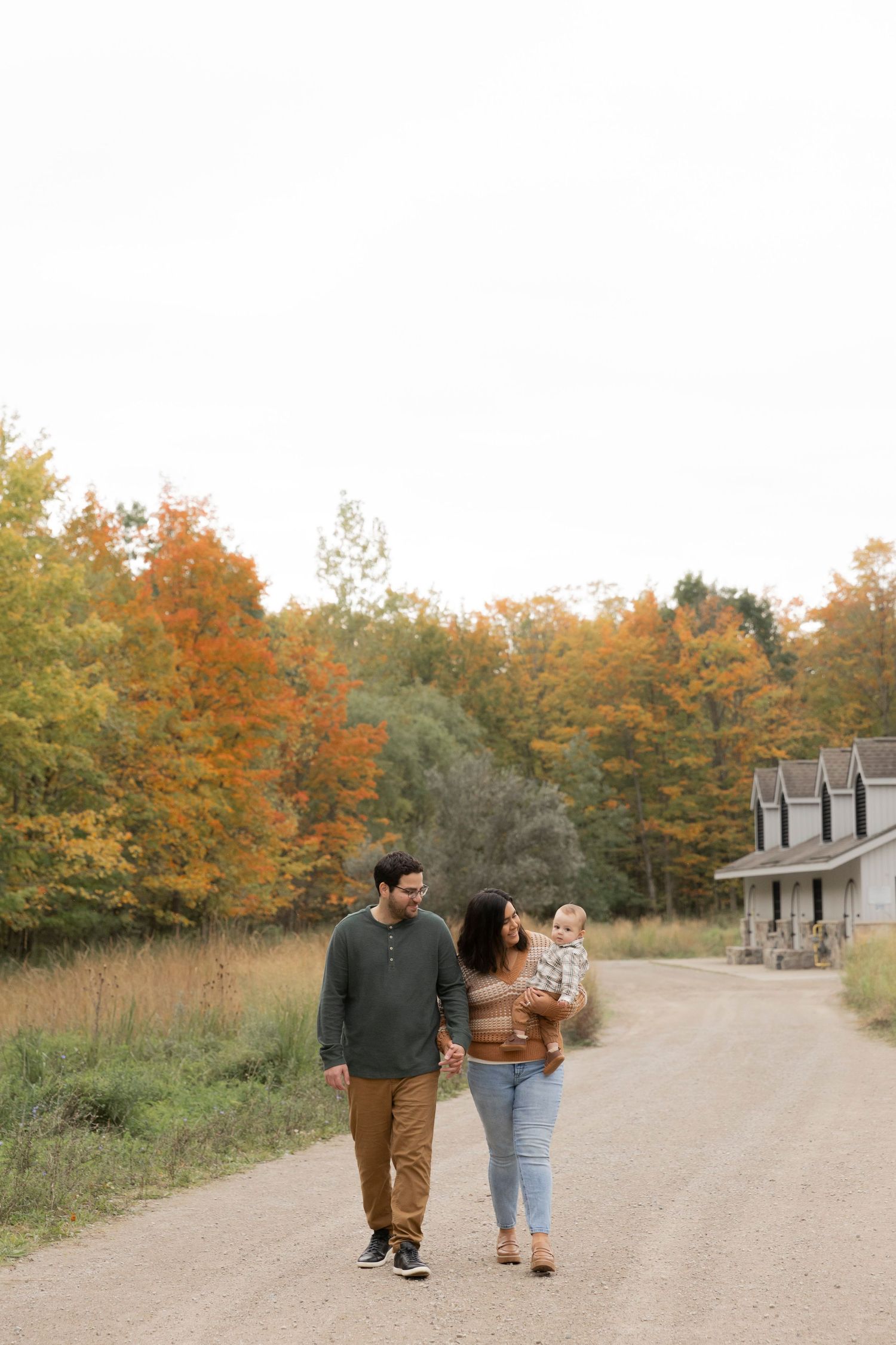 family of 3 walking on a path holding hands and smiling at their baby with fall foliage in Michigan