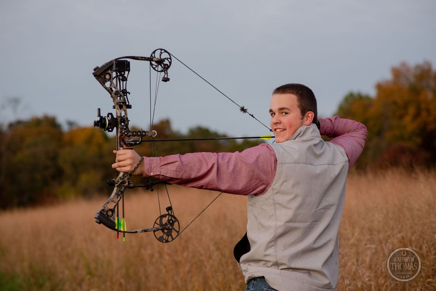Archer draws compound bow during sunset in autumn field.