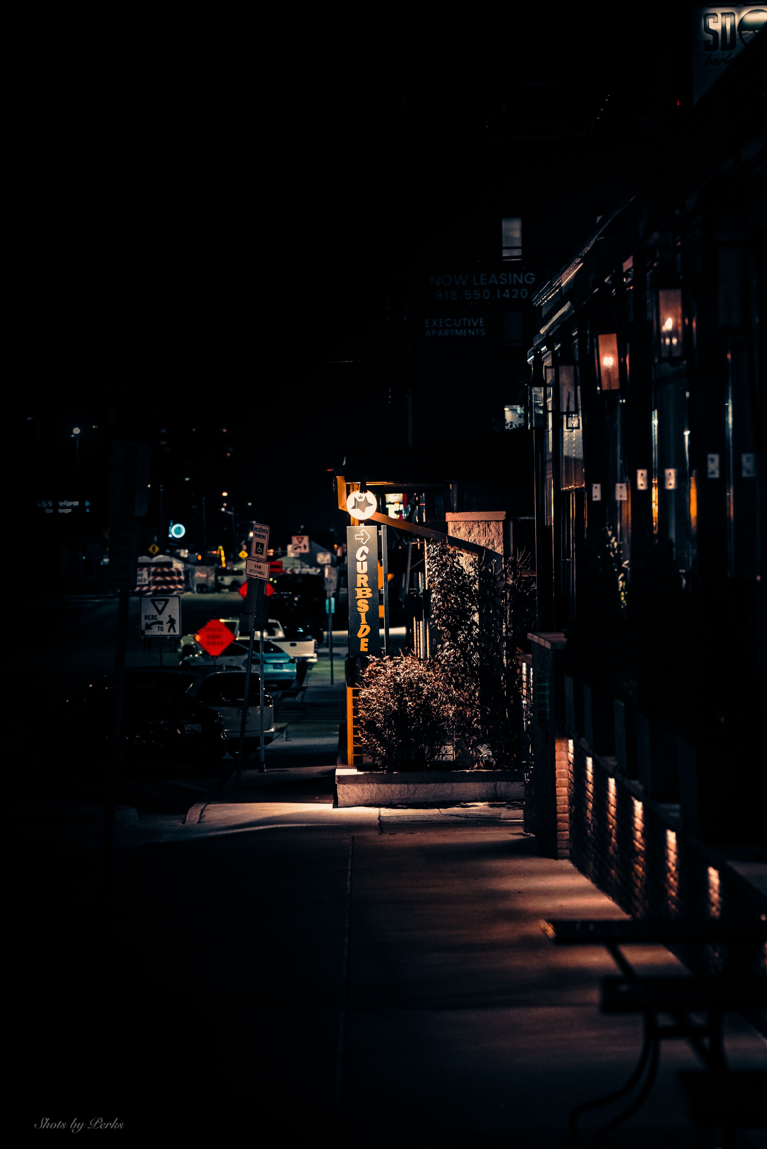 Dark urban street at night illuminated by warm glowing lights and storefronts casting shadows on the sidewalk.