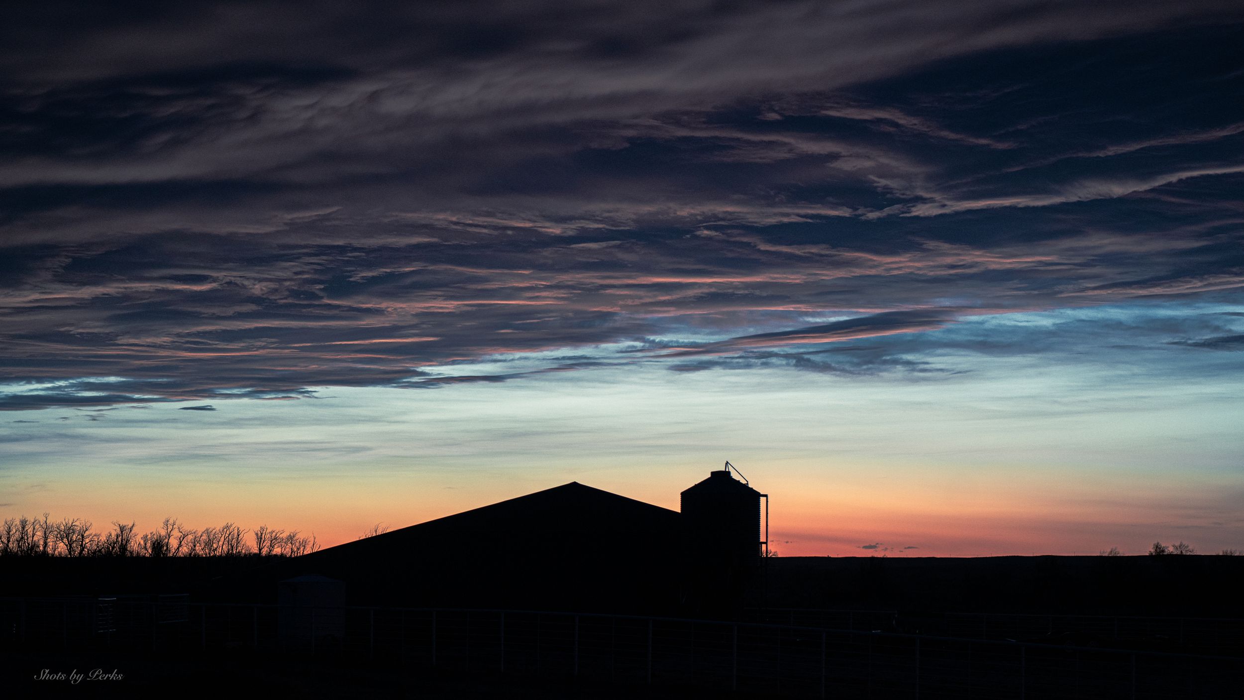 Silhouette of a barn against dramatic sunset sky with dark clouds and orange horizon.