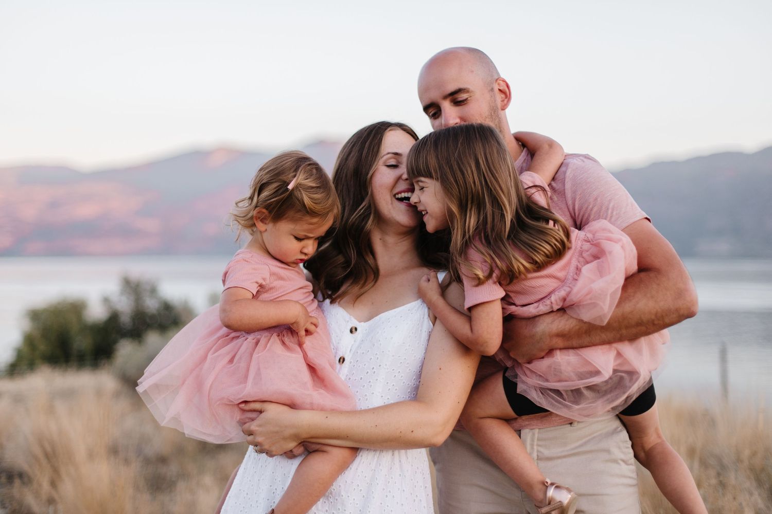 Family of four embracing outdoors at sunset with mountain lake in background.