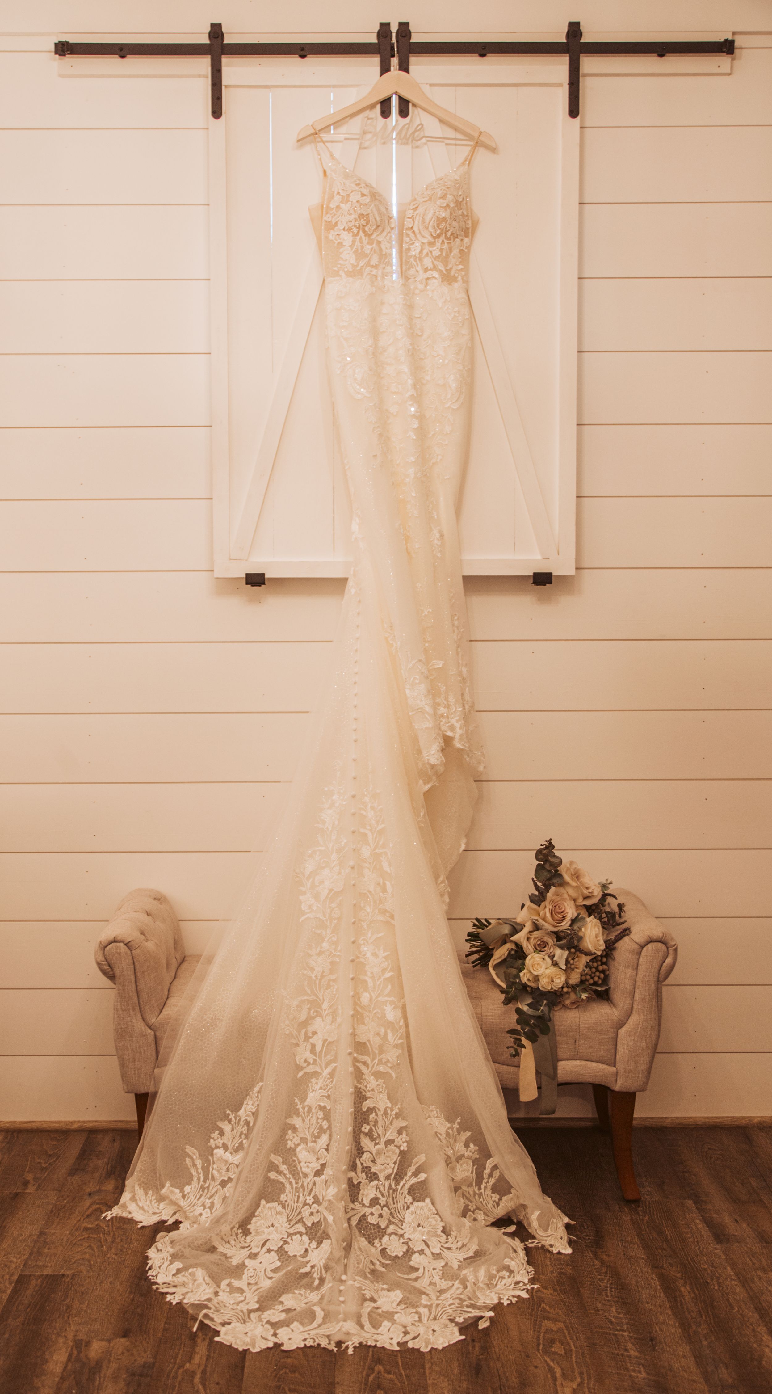 Elegant lace wedding dress hanging on barn door with floral bouquet below.