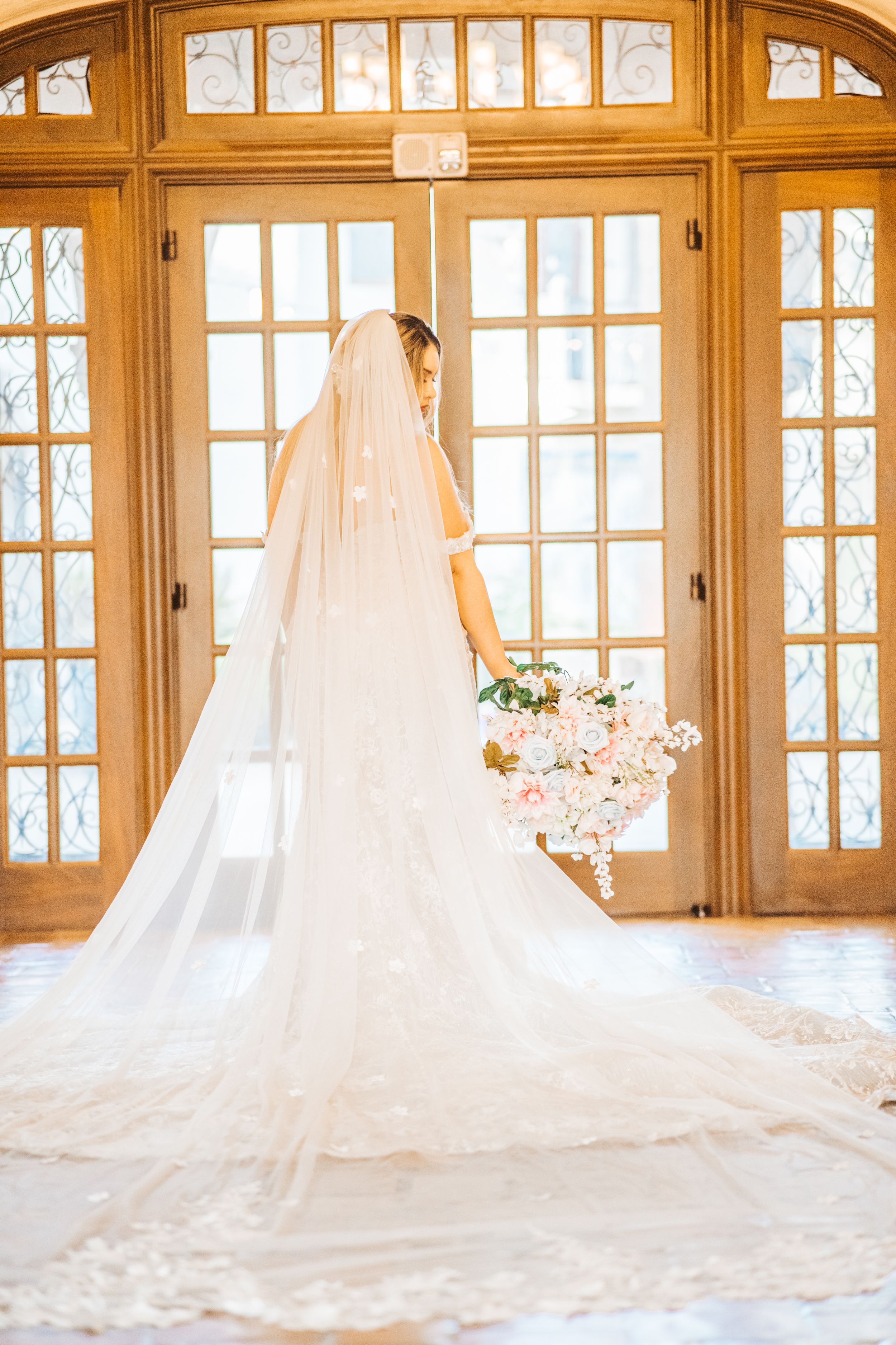 A cathedral-length wedding veil trails behind a bride standing before ornate French doors in natural light.
