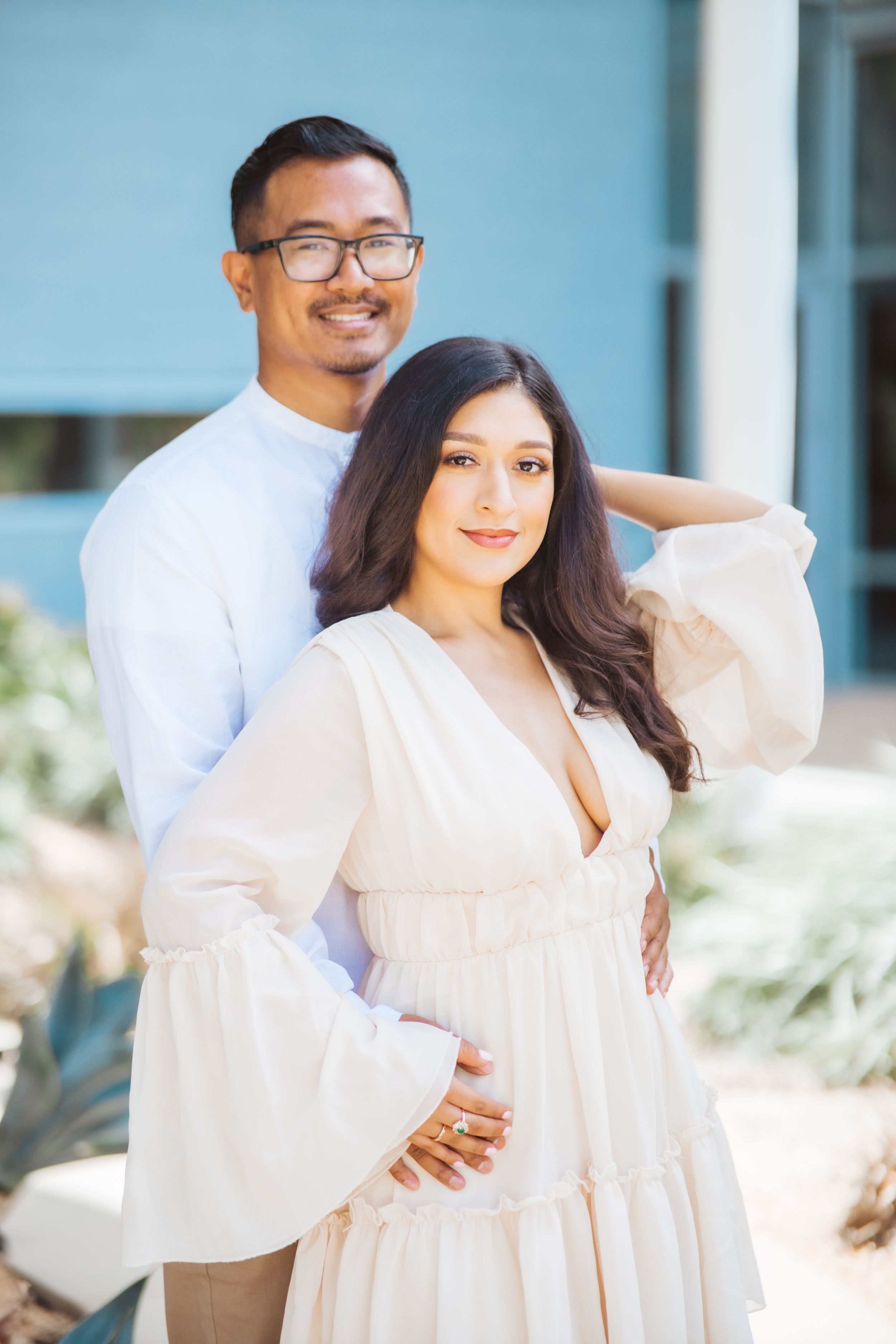 Couple in white outfits posing together against a blue wall background.