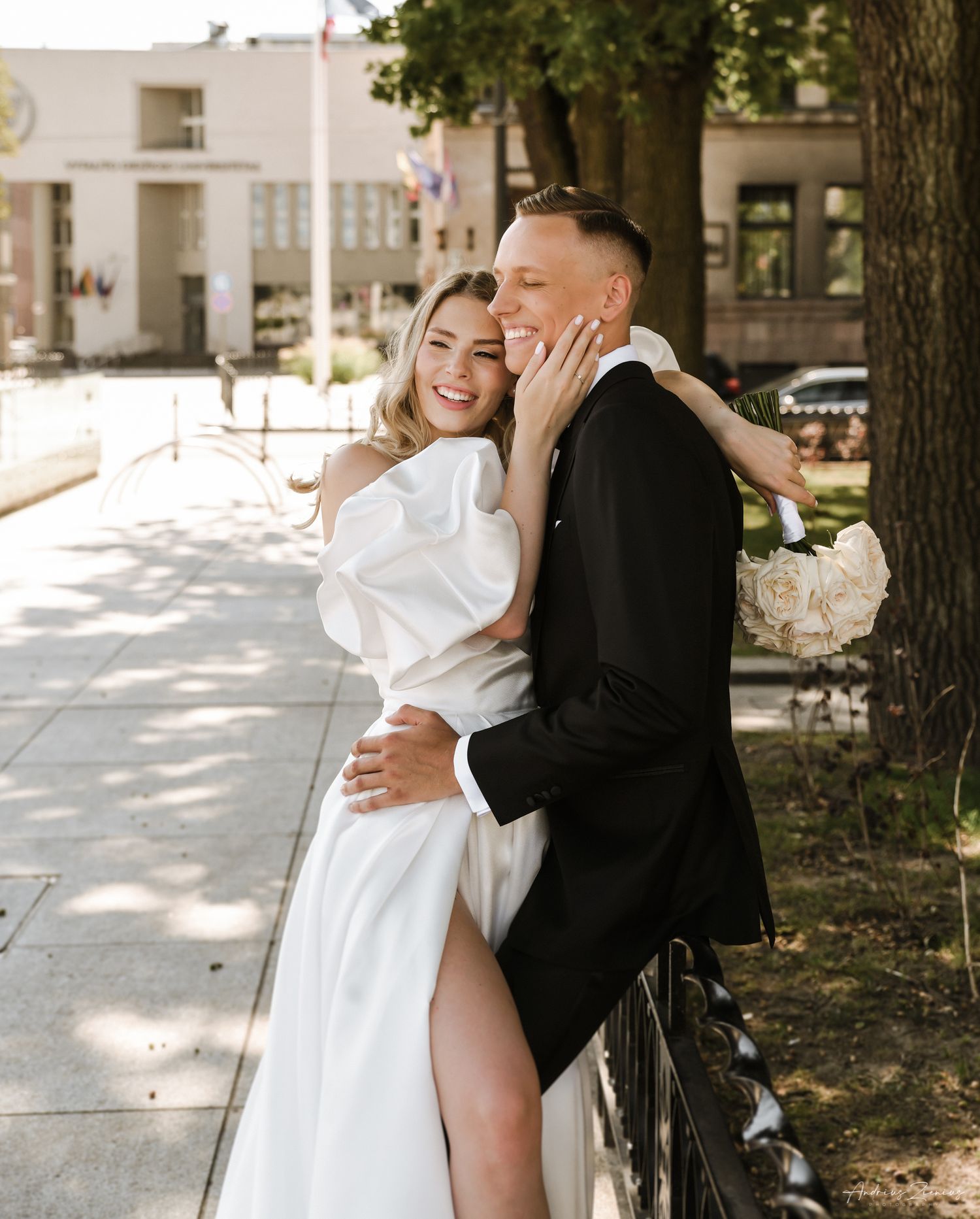 Playful wedding couple embracing on tree-lined sidewalk in summer.