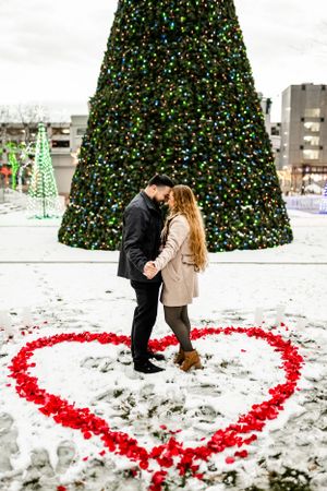 A winter engagement photo with a heart drawn in snow beneath a lit Christmas tree.