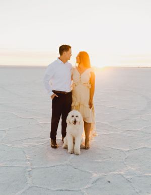 Couple poses with their white fluffy dog on salt flats at golden hour.