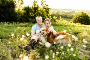 A couple sits in a sunlit meadow with their corgi dog surrounded by wildflowers at sunset.