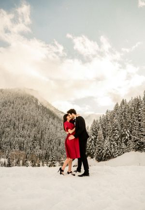 Romantic winter photoshoot in a snow-covered forest landscape with mountains and evergreen trees in the background.