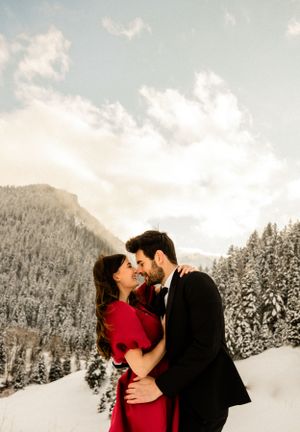 A couple embracing in a snowy mountain landscape with evergreen trees in the background.