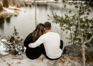 A couple sits close together on a snowy ledge overlooking a serene lake.