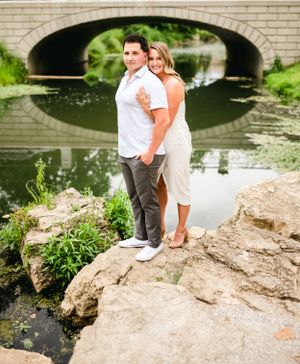 A couple poses on rocky terrain by a stone bridge over calm water.