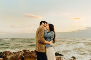 A romantic couple embracing on a rocky beach during sunset with waves crashing in the background.
