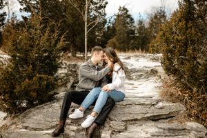 A couple sits together on rocky terrain surrounded by pine trees in winter.