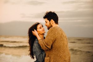 A couple in warm sweaters sharing intimate moments during a sunset beach photoshoot.