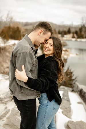 A couple embraces and laughs together in a snowy winter setting by a lake.