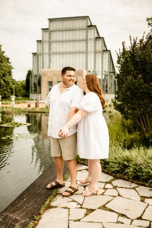 Couple in white outfits embracing near stone path by waterfront building.
