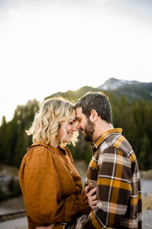 A couple shares an intimate moment at sunset with mountains and pine forest in the background.