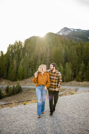 A couple walks together on a gravel path with sunlit mountain peaks and evergreen forest behind them.