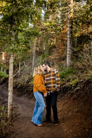 A couple in casual fall attire playfully walking on a wooded dirt trail surrounded by evergreen trees.