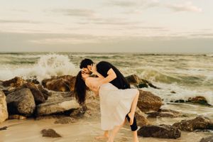 A romantic photoshoot of a couple on a rocky coastline with crashing waves.