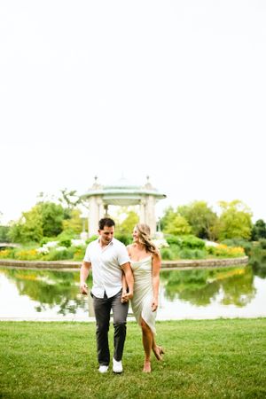 A couple walks together on manicured grass near a gazebo and reflecting pond.