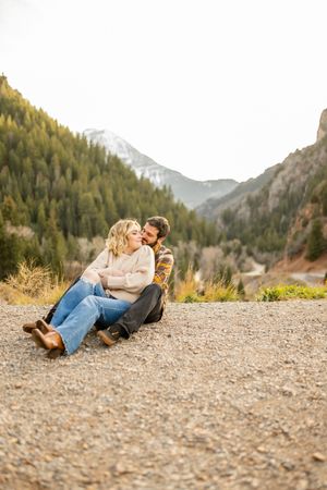 A couple relaxes on gravel ground with mountain views and pine forests in the background.