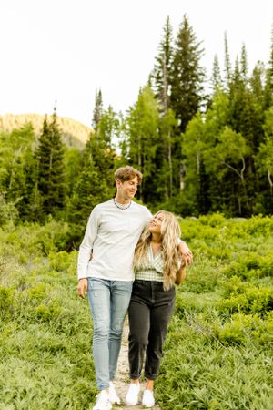 Couple walking and laughing together in a lush mountain meadow with evergreen forest backdrop.