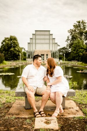 Couple in casual white outfits sitting together near lakeside building.