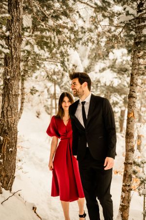 Couple in formal attire walking through snowy pine forest during winter photo session.