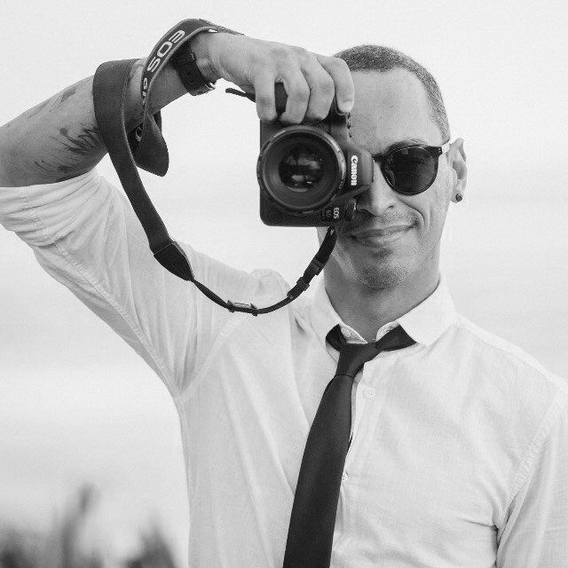 Professional photographer in white shirt and tie holding DSLR camera in black and white portrait.