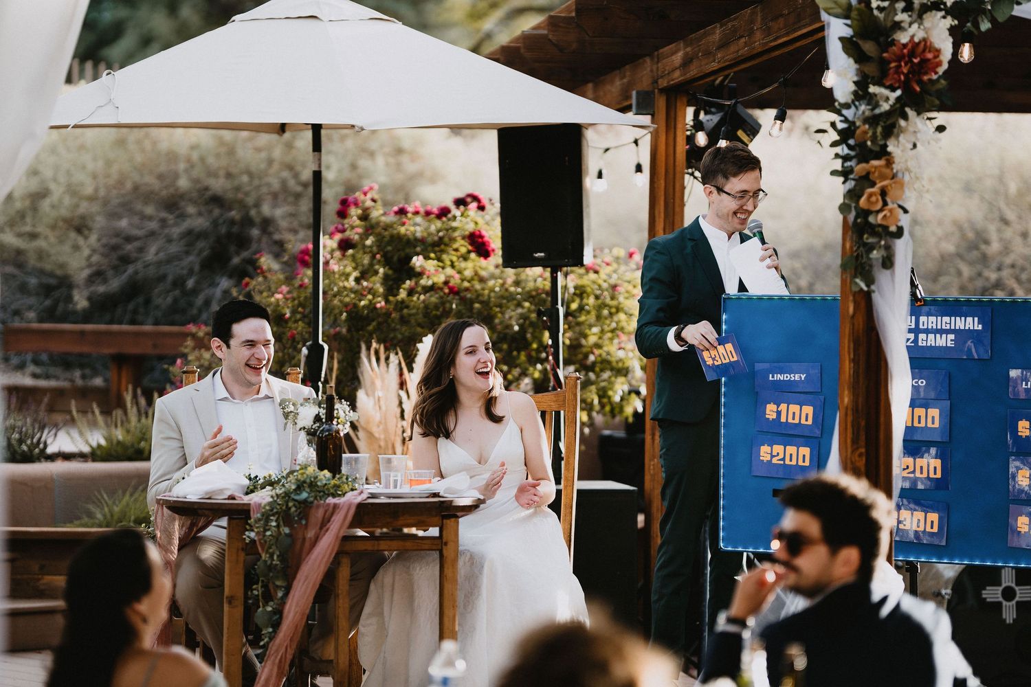 Bride and groom laugh during outdoor wedding reception as guest gives a fun game show speech.cinematic destination wedding photographer