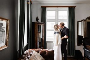 A couple shares an intimate moment by a window with green curtains in an elegantly furnished room.