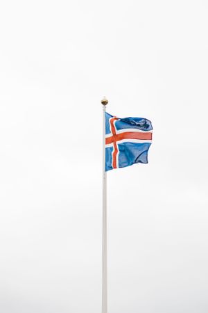 Icelandic flag waving against white cloudy sky on tall flagpole.