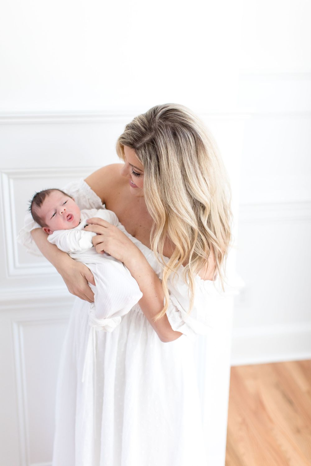 Parent in white dress lovingly cradling and gazing at their newborn baby in a bright room.