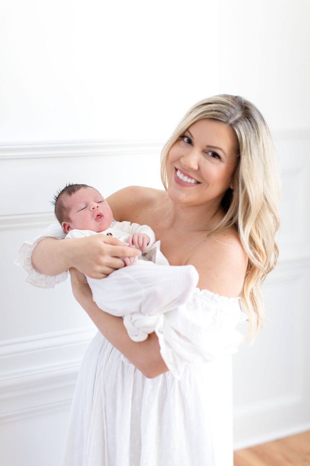 Parent in white dress joyfully holding newborn baby against bright wall.