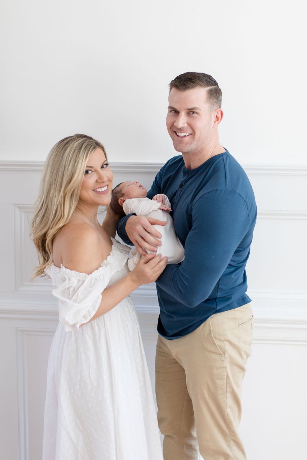 Happy couple in navy and white holding their newborn baby against a white wall.