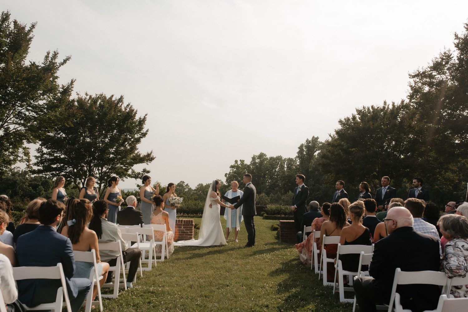 Outdoor wedding ceremony set up with white chairs and guests watching couple exchange vows under trees.