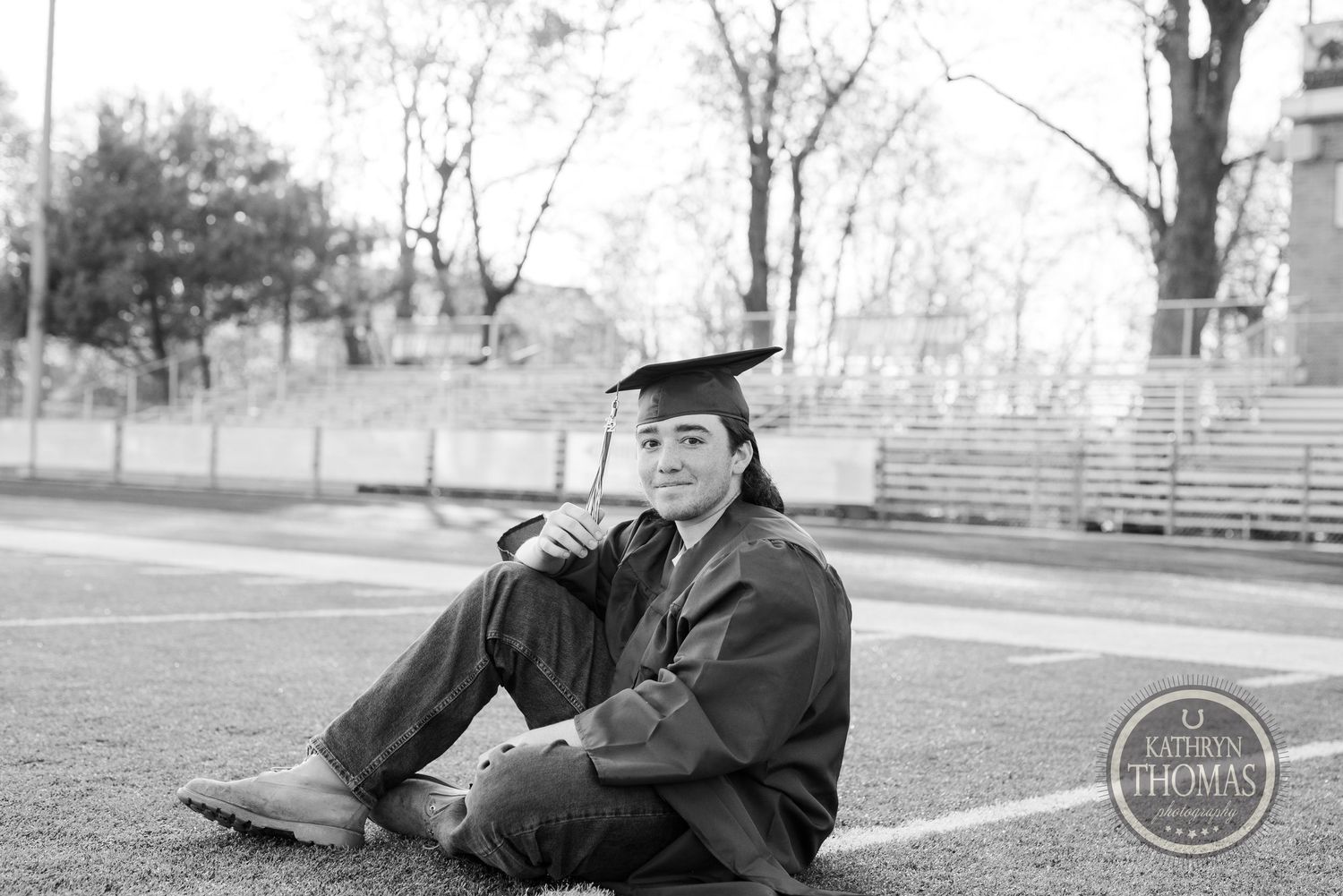 Black and white photo of graduate in cap and gown sitting on football field during photo session.