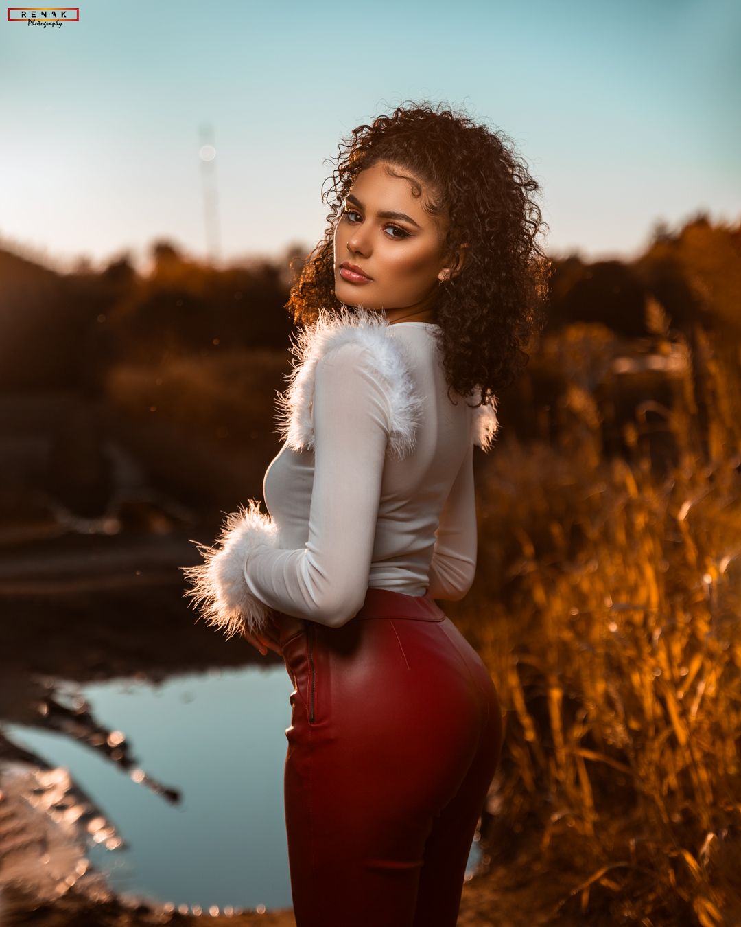 Woman in white top and red skirt poses at sunset near water with natural curly hair.