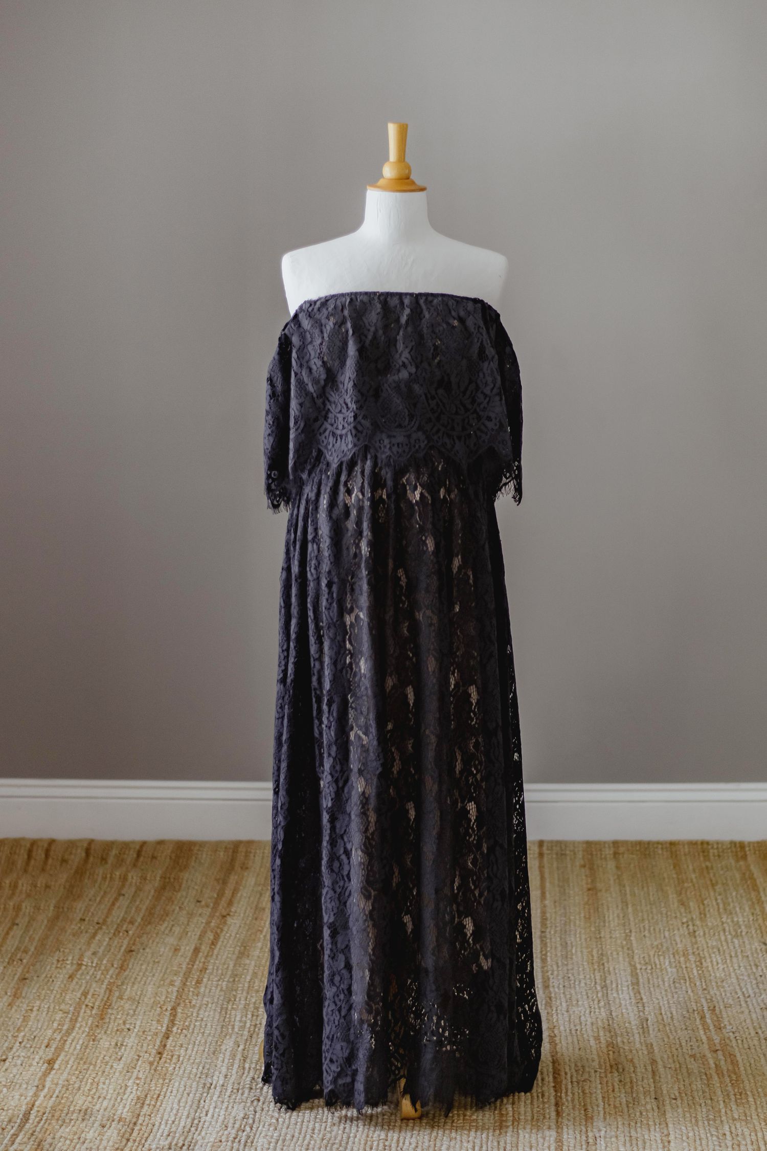 Black lace off-shoulder maternity gown displayed on mannequin against grey wall.