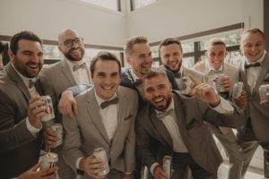 Groomsmen in bow ties and suits smile while holding drinks for a group photo.