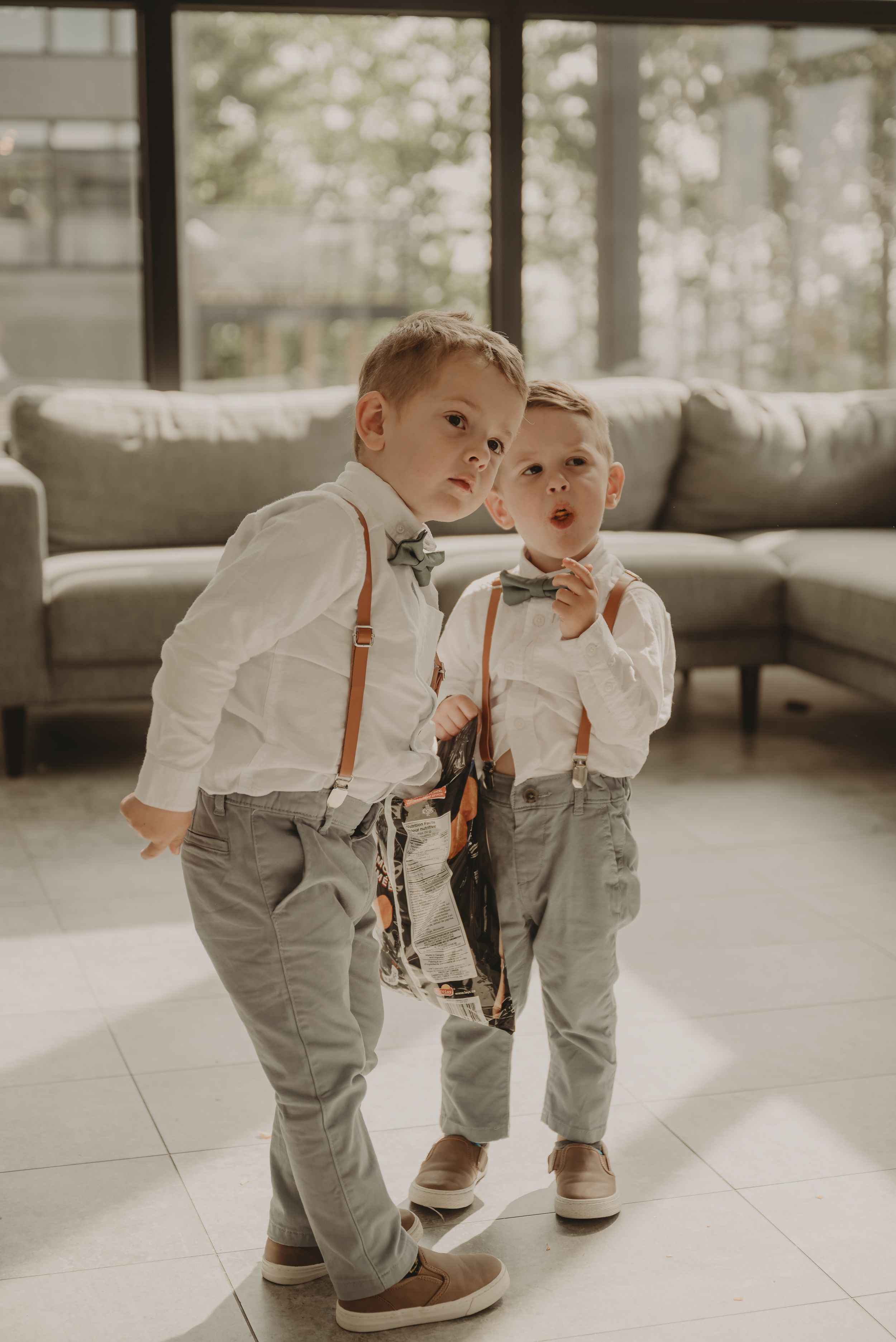 Two young children in matching suspenders and white shirts pose together in a living room with modern decor.
