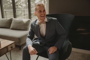 Groom seated in gray armchair laughing during wedding preparations.