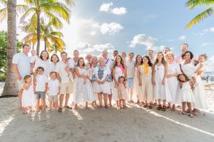 Large group portrait of people in white and beige clothing on a tropical beach with palm trees.