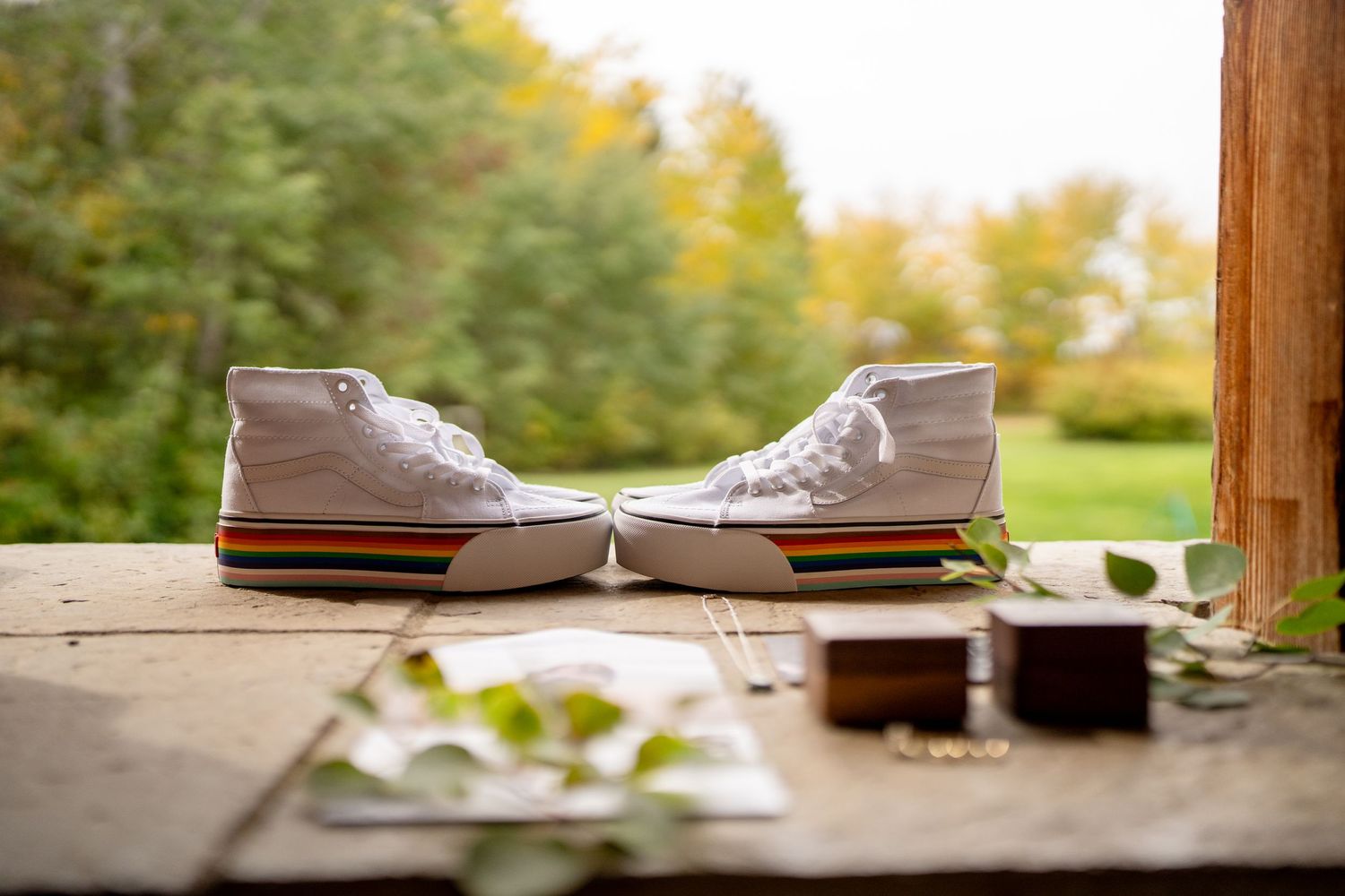 White canvas sneakers arranged with wedding rings on a wooden surface with fall foliage in background.